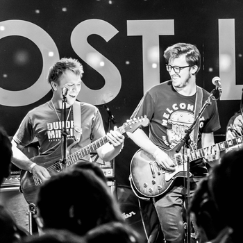 Two guitarists play on stage in front of a crowd, with a large sign in the background containing the letters "OSTL." Both musicians are focused on their performance.