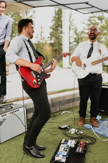 Two musicians, one playing a red electric guitar and the other playing a white bass guitar, perform outdoors on a grassy area with equipment and amplifiers nearby. Both are smiling and dressed in formal attire.