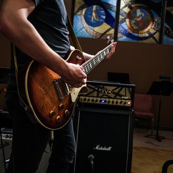 A person plays an electric guitar in front of an amplifier in a room with stained glass windows in the background.