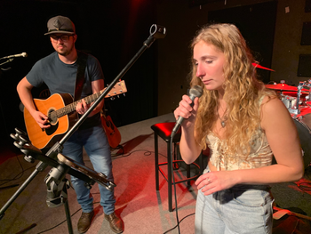Two musicians perform on stage, one playing an acoustic guitar and the other singing into a microphone, with music stands and equipment visible in the background.