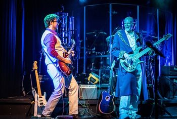 Two musicians perform on stage with guitars. One is dressed in a light-colored outfit, the other in a dark coat with a ceremonial chain. They appear to be enjoying the performance.